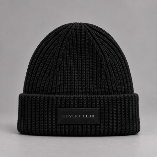 A black ribbed knit beanie hat with a folded cuff and a small black label reading 'COVERT CLUB' in white text, photographed against a neutral gray background.